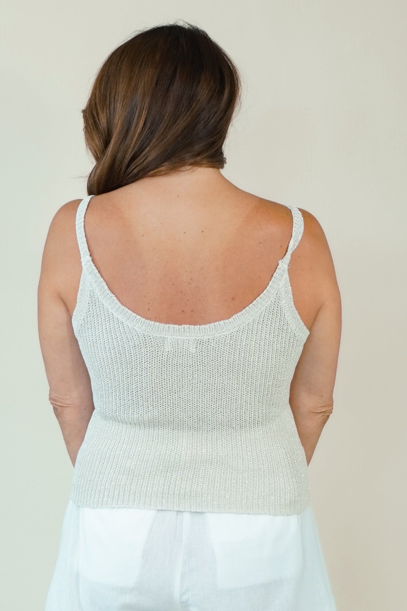 A woman viewed from the back wearing a light cream ribbed knit camisole top with thin straps and a scoop neckline, paired with white bottoms, against a plain light beige background.