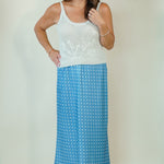 A woman modeling a two-piece outfit consisting of a white sleeveless knit top with floral embroidery and a blue maxi skirt with a white polka dot pattern, set against a plain beige studio background.