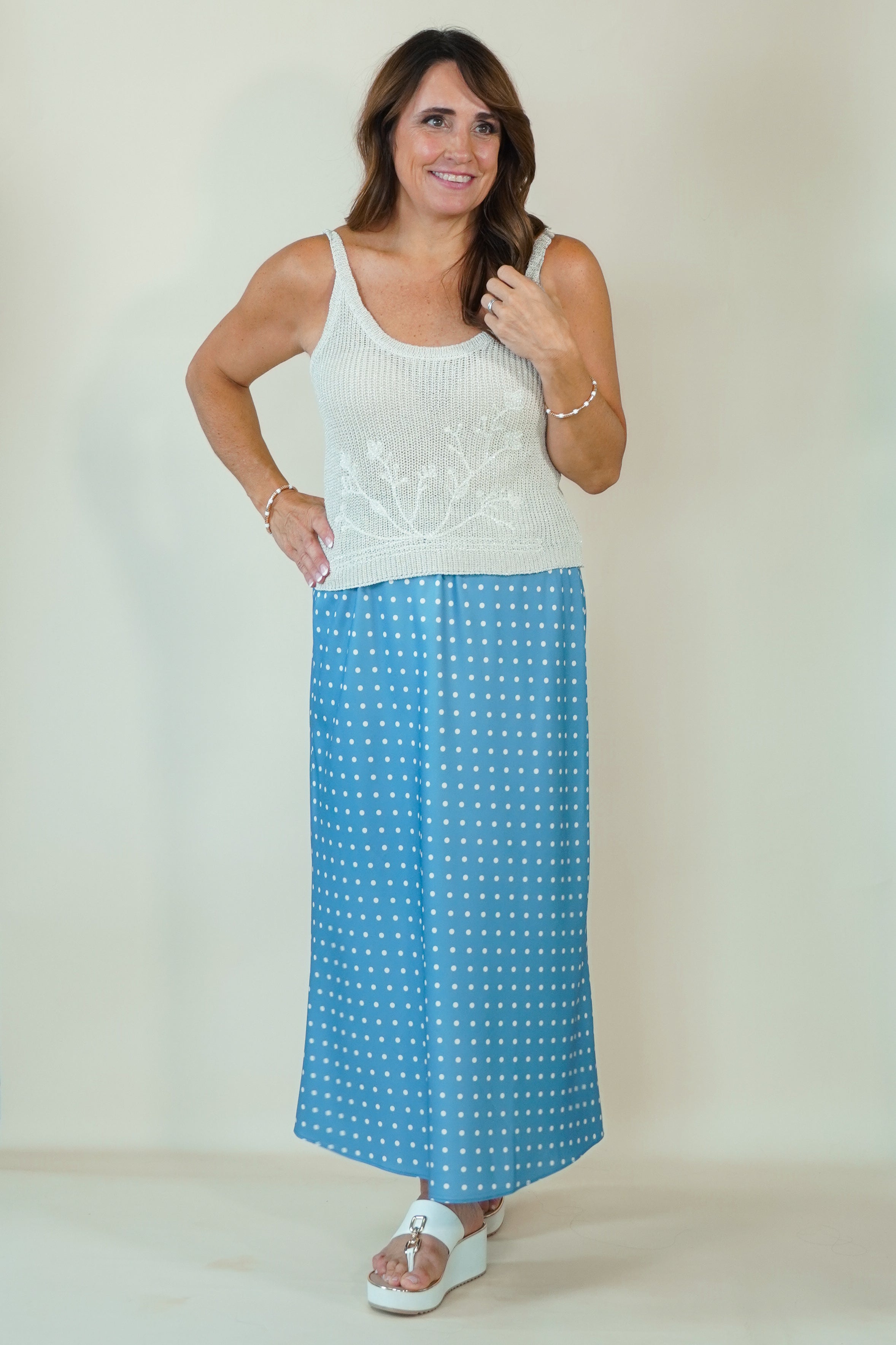 A woman modeling a two-piece outfit consisting of a white sleeveless knit top with floral embroidery and a blue maxi skirt with a white polka dot pattern, set against a plain beige studio background.