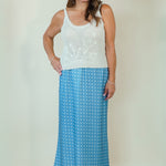 A woman modeling a two-piece outfit consisting of a white sleeveless knit top with a floral pattern and a blue maxi skirt with white polka dots, standing against a plain beige studio backdrop.