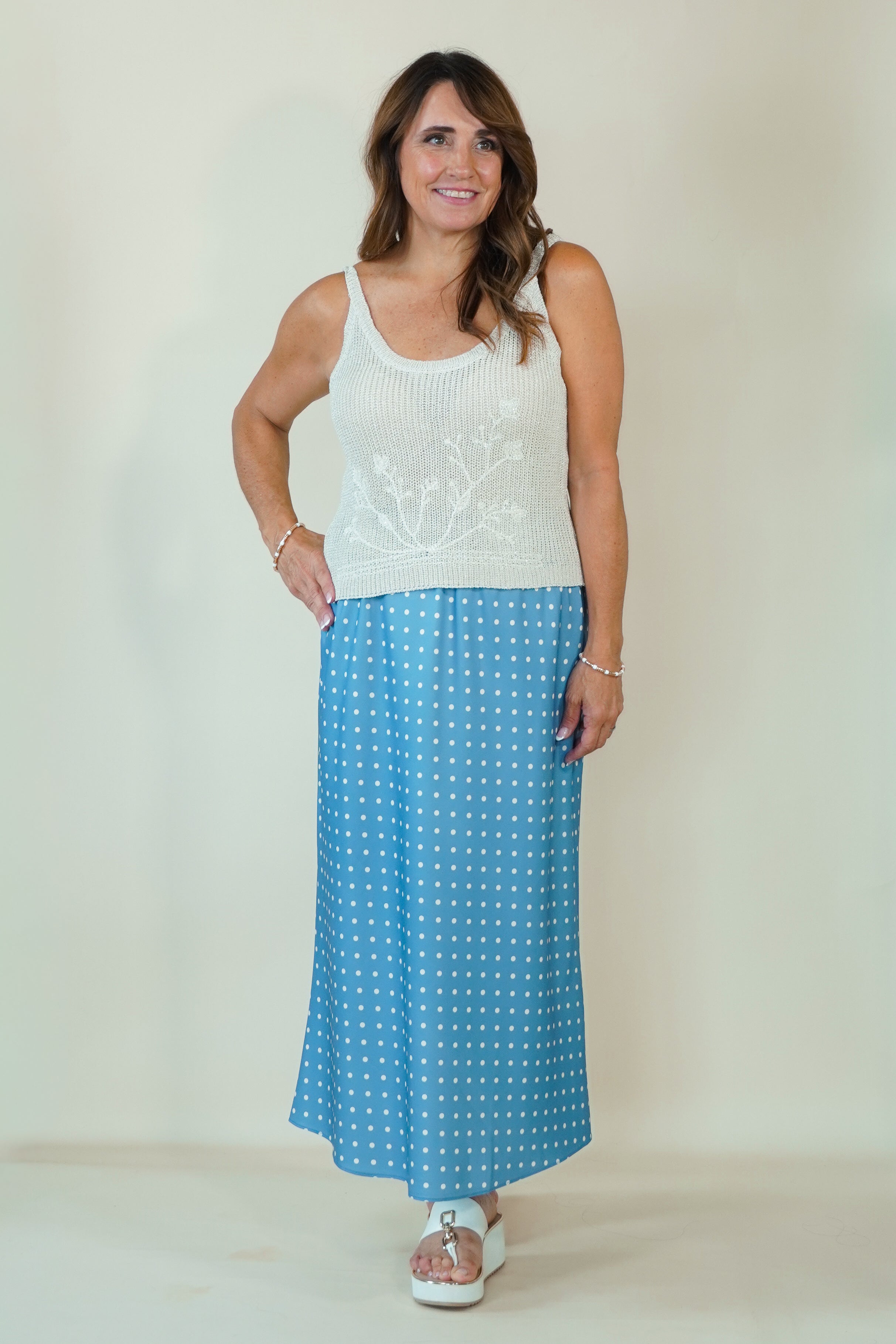 A woman modeling a two-piece outfit consisting of a white sleeveless knit top with a floral pattern and a blue maxi skirt with white polka dots, standing against a plain beige studio backdrop.