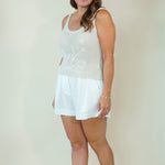 A woman modeling a white sleeveless knit top with floral embroidery and white shorts, standing against a plain beige studio background.