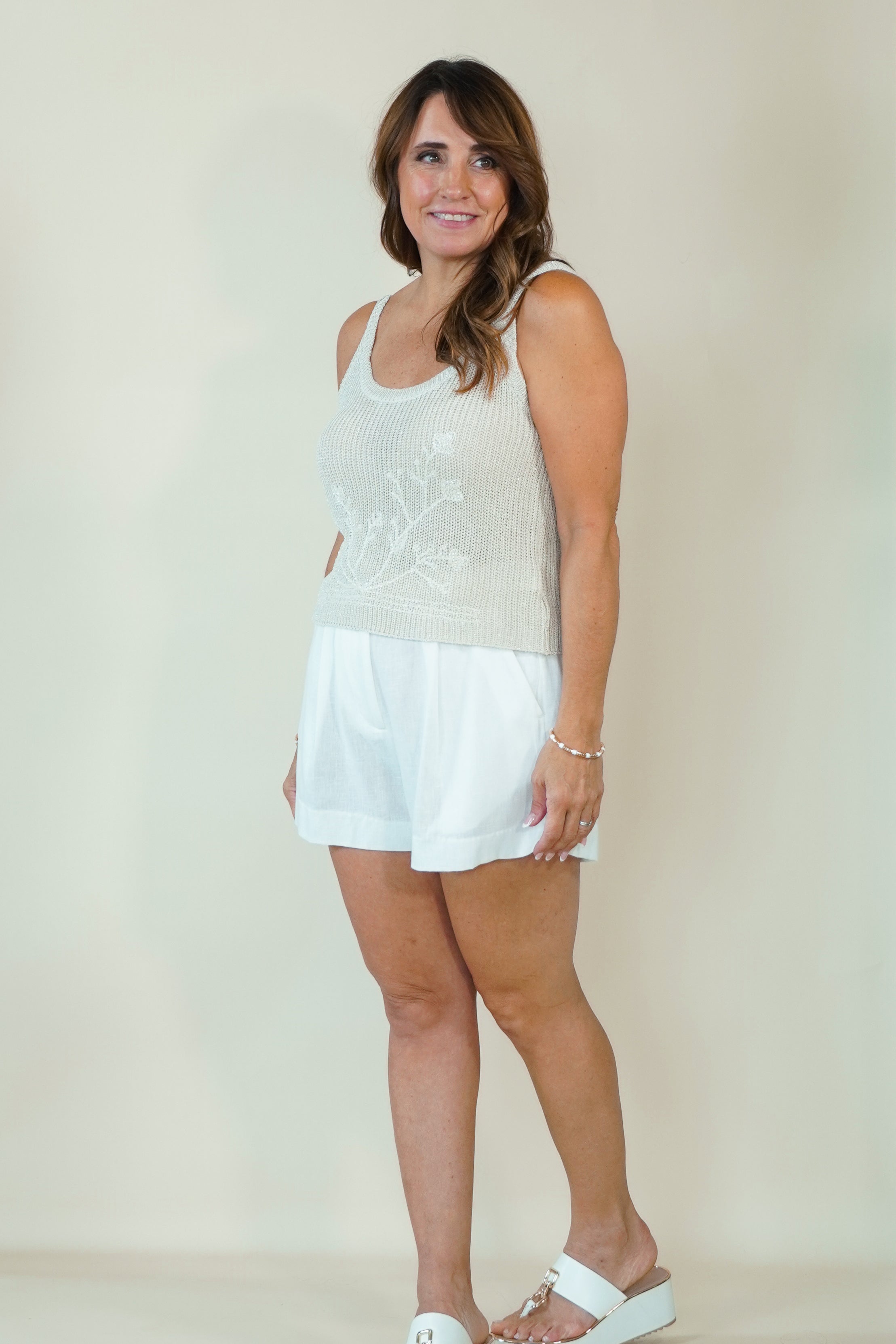 A woman modeling a white sleeveless knit top with floral embroidery and white shorts, standing against a plain beige studio background.