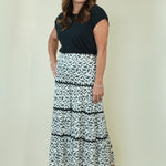 A woman modeling a black short-sleeve top and a long tiered maxi skirt with a black and white abstract pattern, standing against a plain cream-colored studio background.