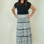 A woman modeling a black short-sleeve top and a black and white patterned tiered maxi skirt, standing against a plain cream-colored studio background.