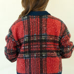 A woman viewed from behind wearing a red and black plaid knit jacket with dark navy ribbed trim, paired with blue jeans and brown heels, against a plain off-white background.
