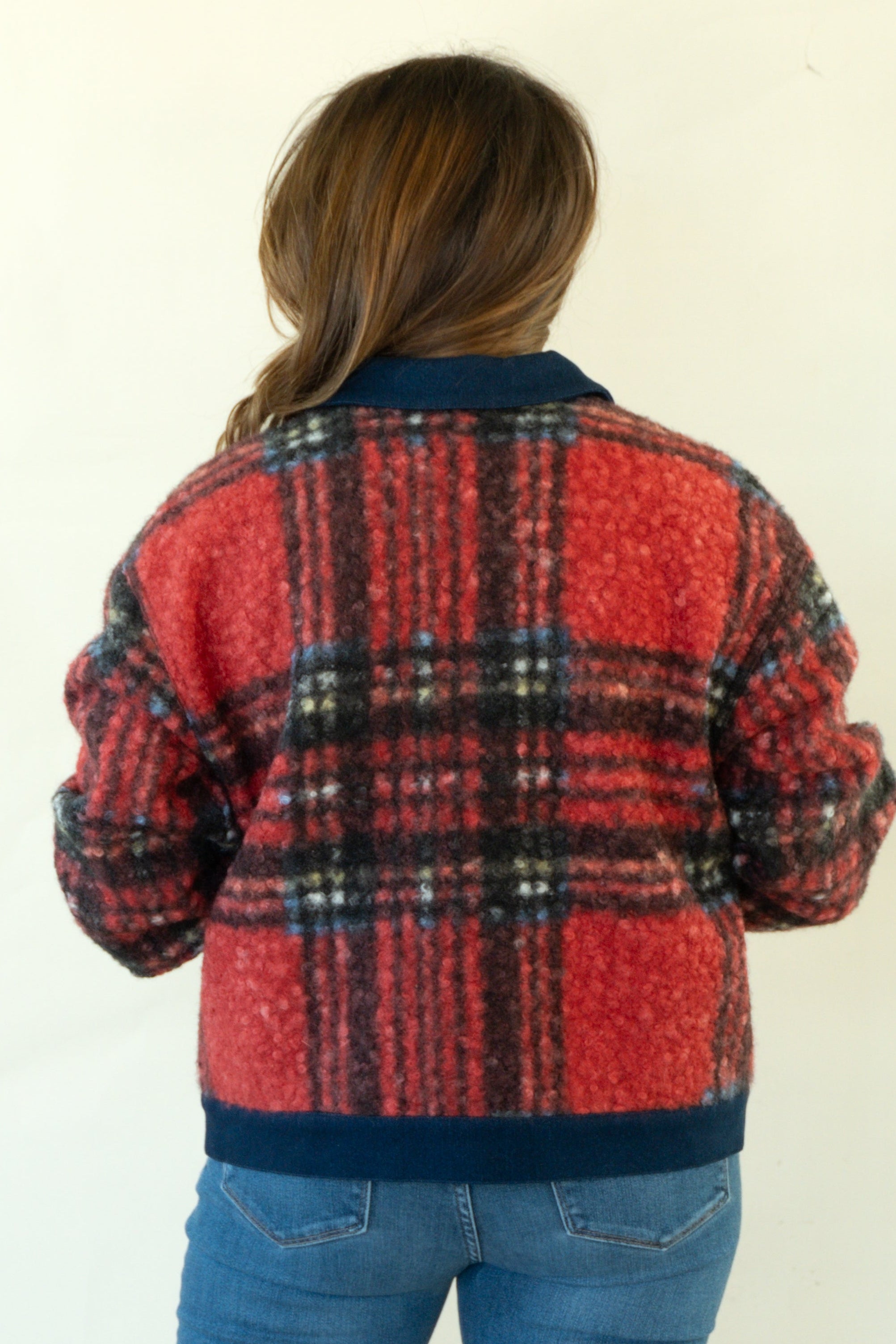 A woman viewed from behind wearing a red and black plaid knit jacket with dark navy ribbed trim, paired with blue jeans and brown heels, against a plain off-white background.