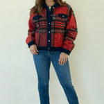 A woman modeling a red and navy plaid button-up jacket with blue jeans and tan ankle boots, standing against a plain off-white studio background.