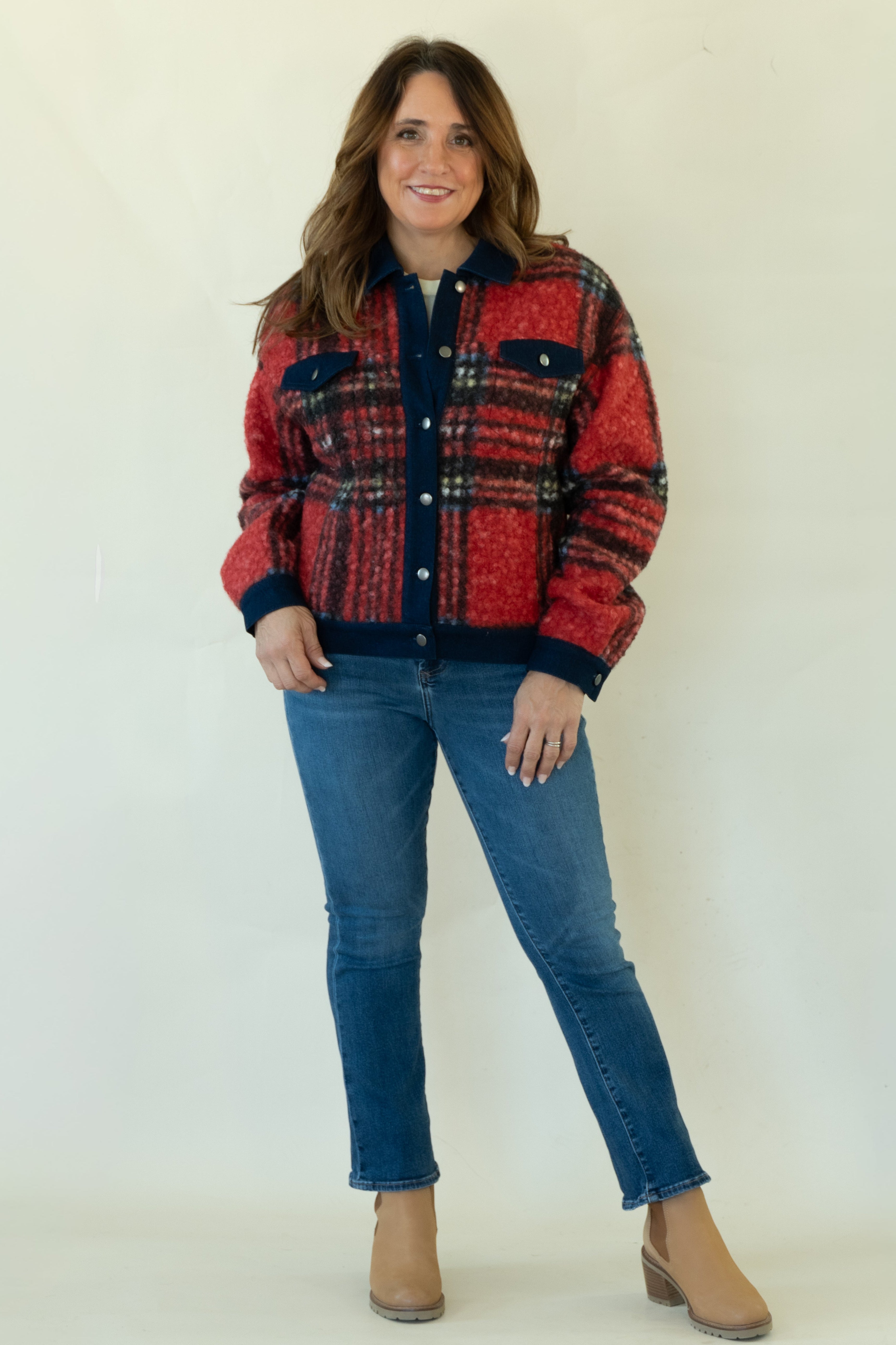 A woman modeling a red and navy plaid button-up jacket with blue jeans and tan ankle boots, standing against a plain off-white studio background.