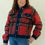 A woman modeling a red and navy plaid button-up jacket with blue jeans and tan ankle boots, standing against a plain off-white studio background.