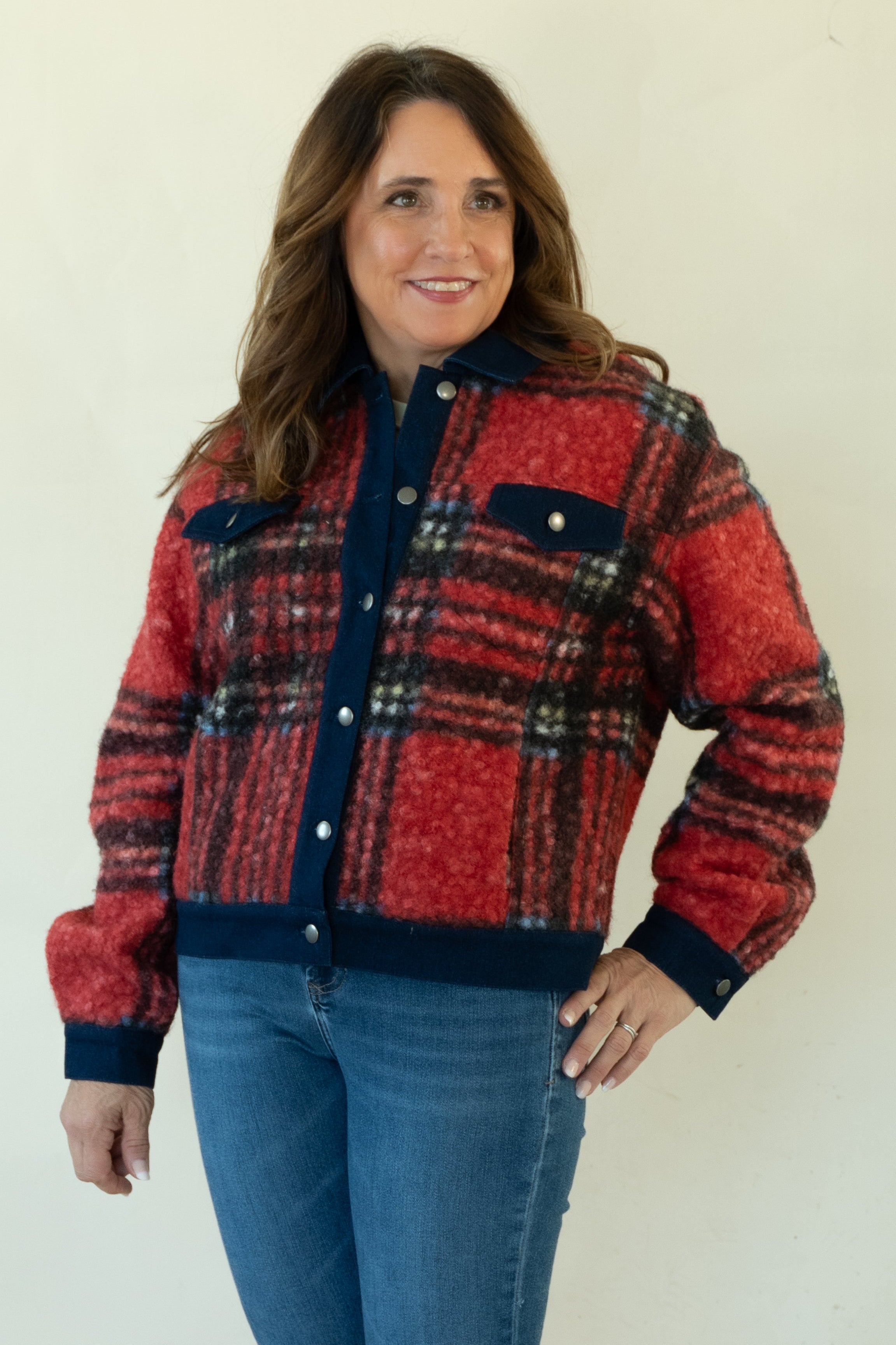 A woman modeling a red and navy plaid button-up jacket with blue jeans and tan ankle boots, standing against a plain off-white studio background.