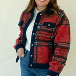 A woman modeling a red and navy plaid button-up jacket with a white top, blue jeans, and tan ankle boots, standing against a plain white studio background.