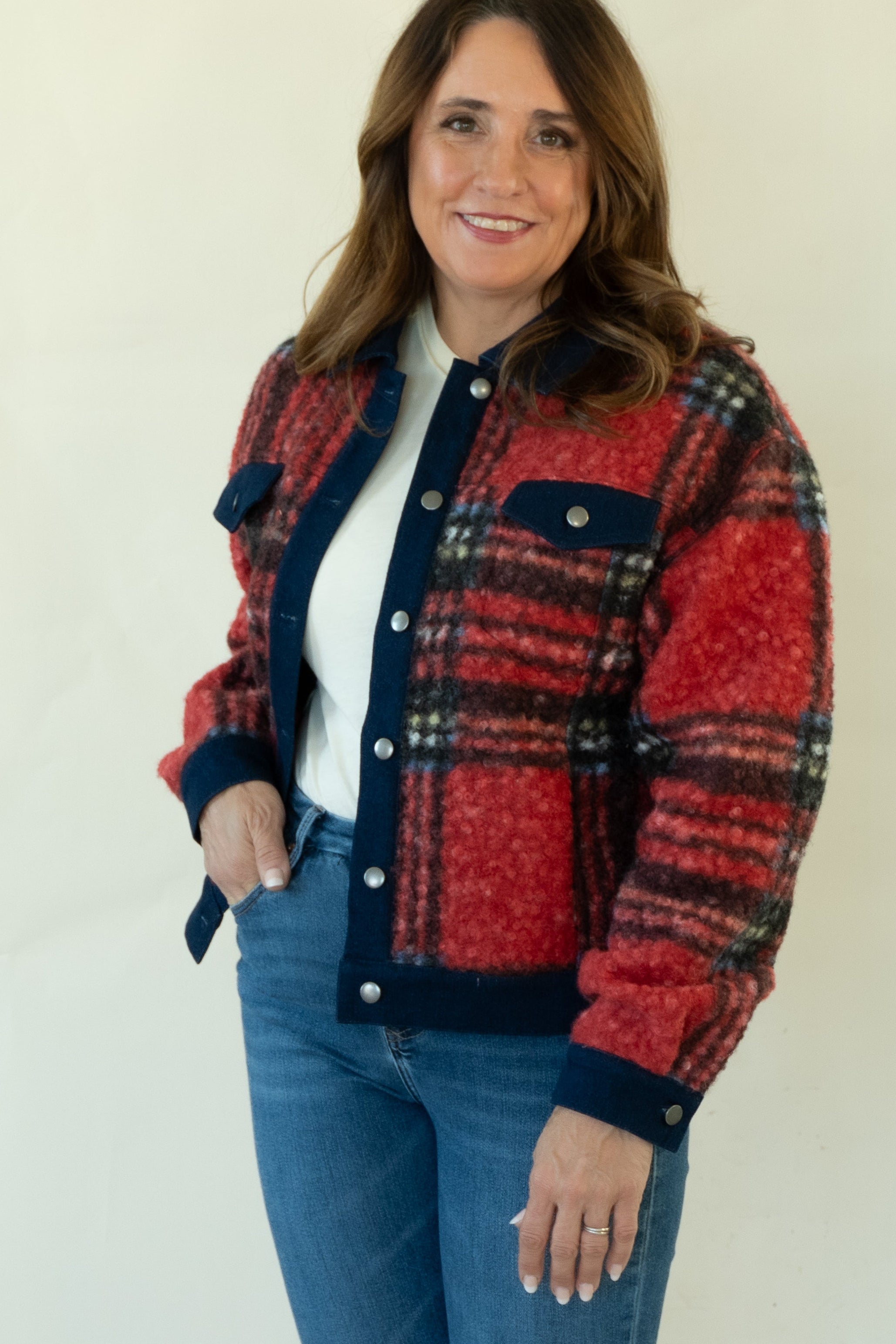 A woman modeling a red and navy plaid button-up jacket with a white top, blue jeans, and tan ankle boots, standing against a plain white studio background.