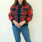 A woman modeling a red and navy plaid button-up jacket with blue jeans and tan ankle boots, standing against a plain white studio background.