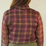 A woman shown from the back wearing a rust-brown and pink plaid long-sleeve shirt, an olive green skirt, and beige wedge sandals, standing against a plain off-white background.