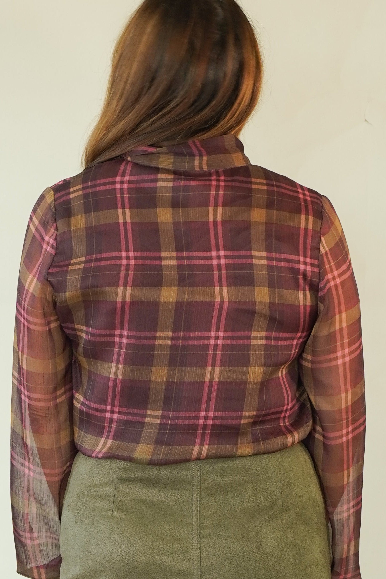 A woman shown from the back wearing a rust-brown and pink plaid long-sleeve shirt, an olive green skirt, and beige wedge sandals, standing against a plain off-white background.
