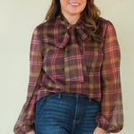 A woman modeling a long-sleeve plaid blouse with a tie-neck detail and blue jeans, standing against a plain light beige studio background.