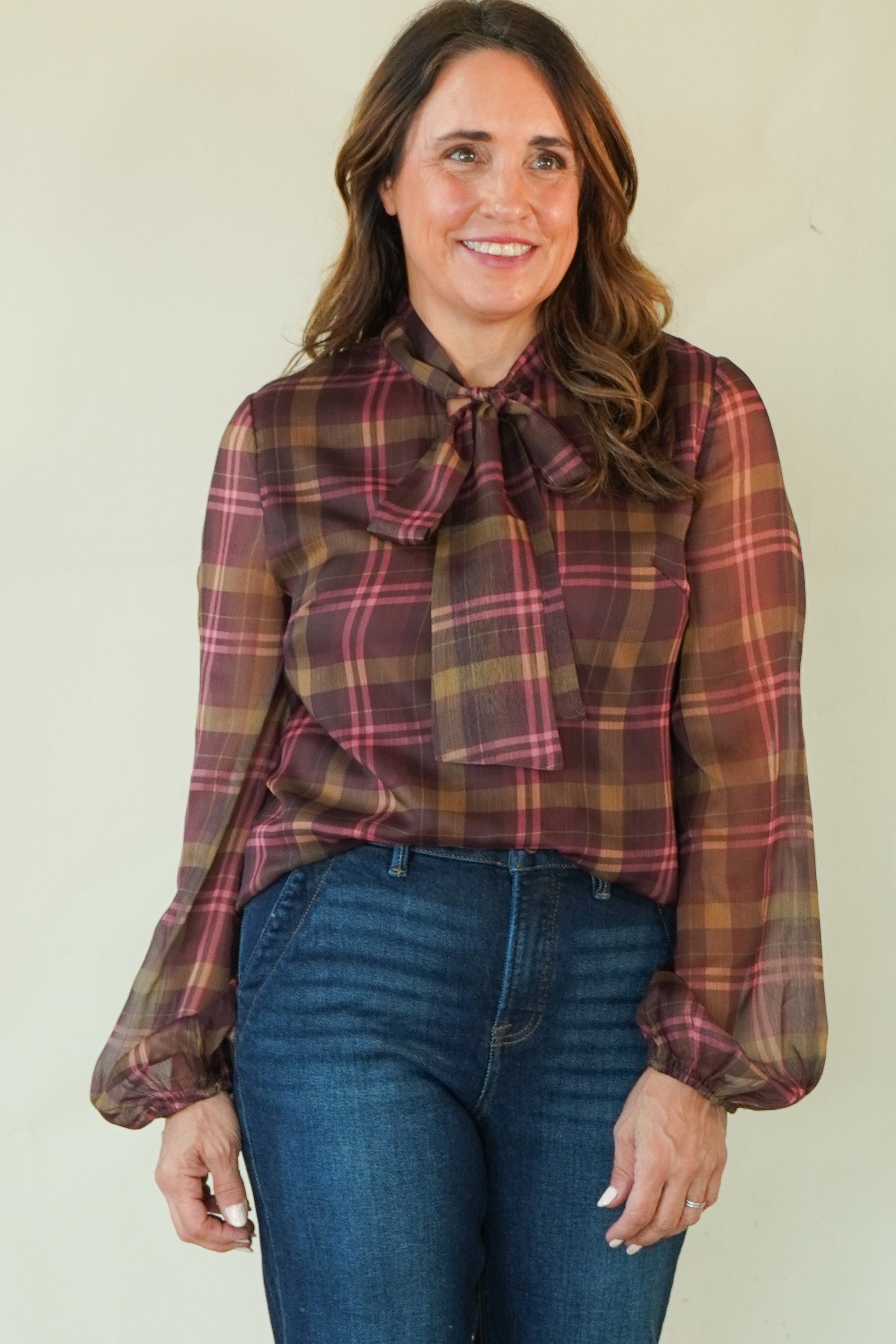 A woman modeling a long-sleeve plaid blouse with a tie-neck detail and blue jeans, standing against a plain light beige studio background.