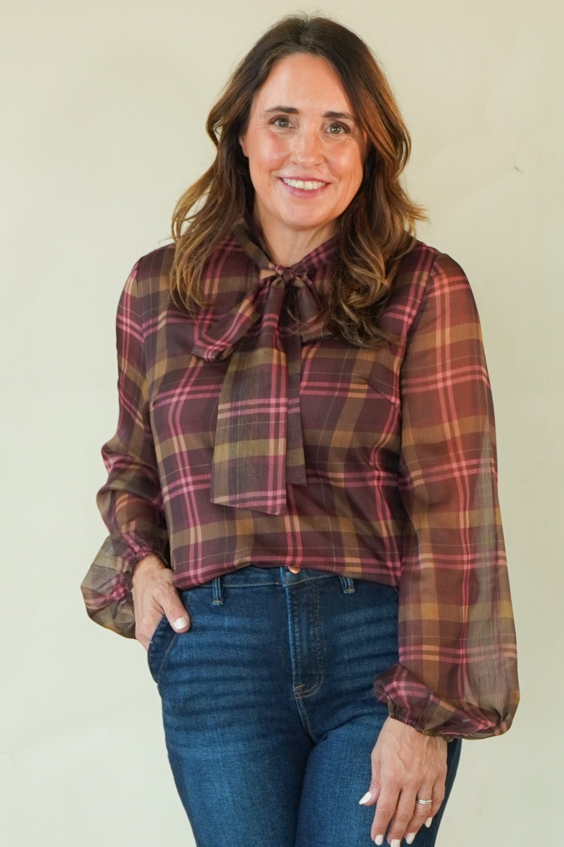 A woman modeling a long-sleeve plaid blouse with a tie-neck detail and blue jeans, standing against a plain light beige studio background.