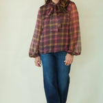A woman modeling a long-sleeve plaid blouse with a bow tie neckline, paired with blue jeans and brown shoes, standing against a plain light beige studio background.