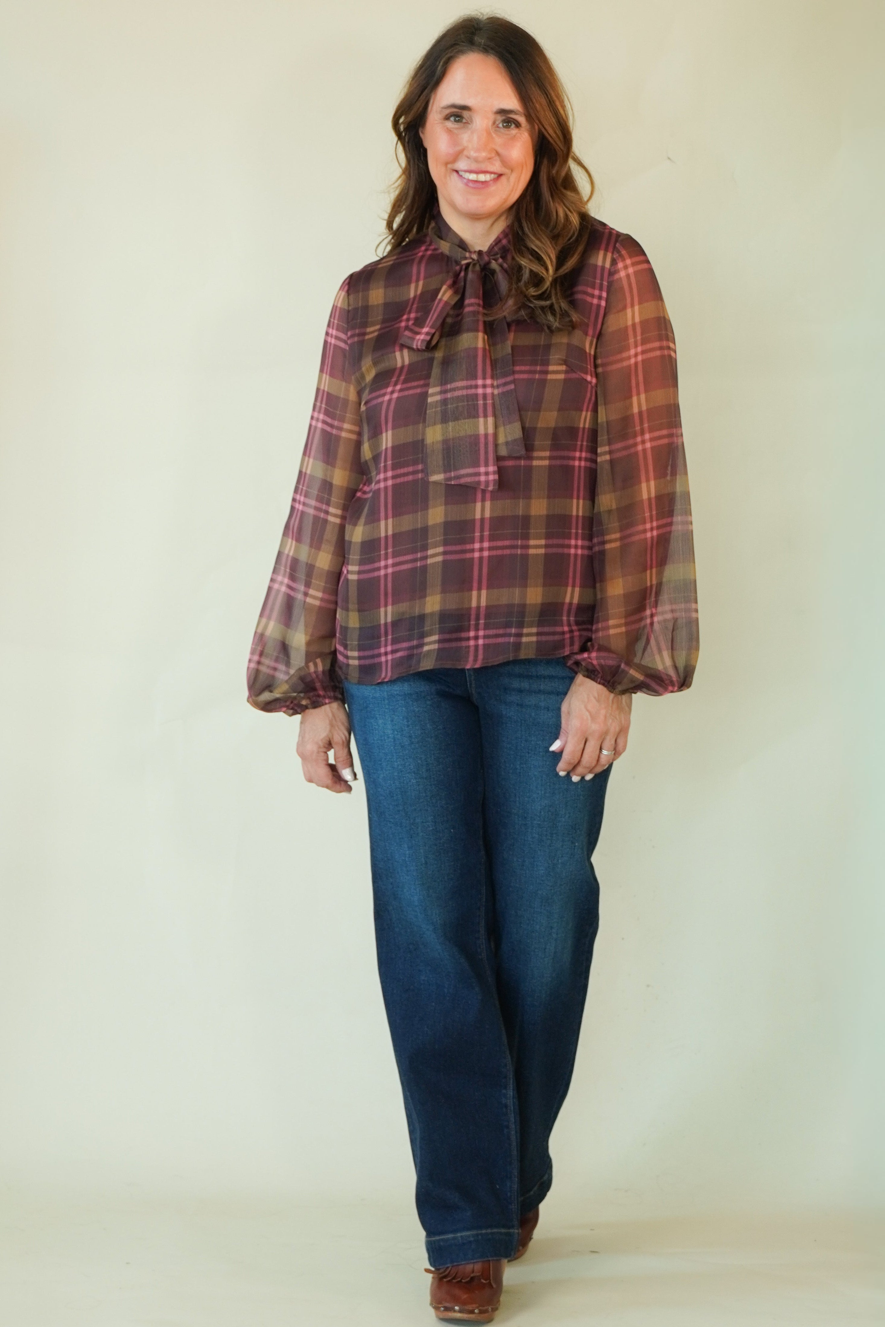 A woman modeling a long-sleeve plaid blouse with a bow tie neckline, paired with blue jeans and brown shoes, standing against a plain light beige studio background.