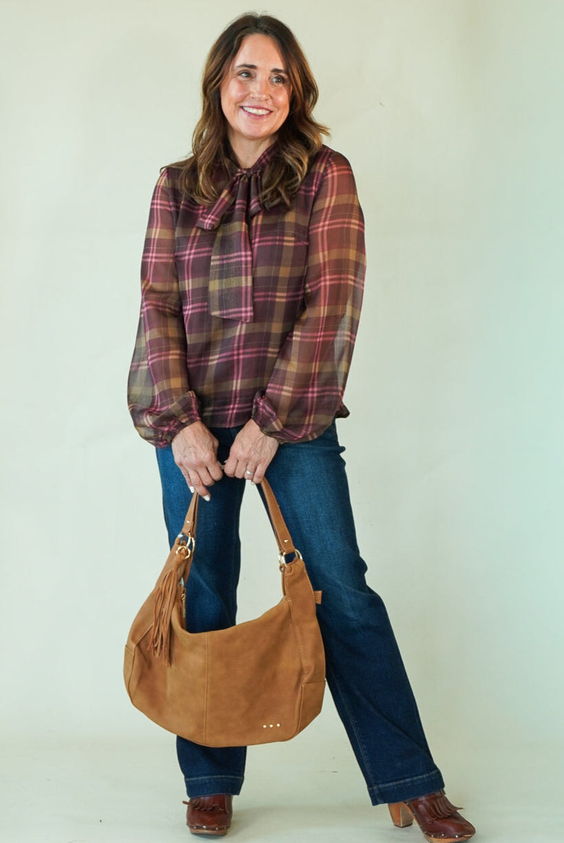 A woman modeling a brown and red plaid long-sleeve blouse, blue jeans, and brown boots while holding a tan suede hobo bag against a plain light green studio background.