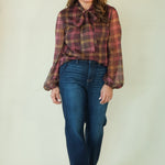 A woman modeling a long-sleeve plaid blouse with a tie-neck detail and blue jeans, standing against a plain light beige studio background.