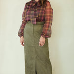 A woman modeling a long-sleeve olive green midi skirt paired with a brown and red plaid blouse, standing against a plain light beige studio background.