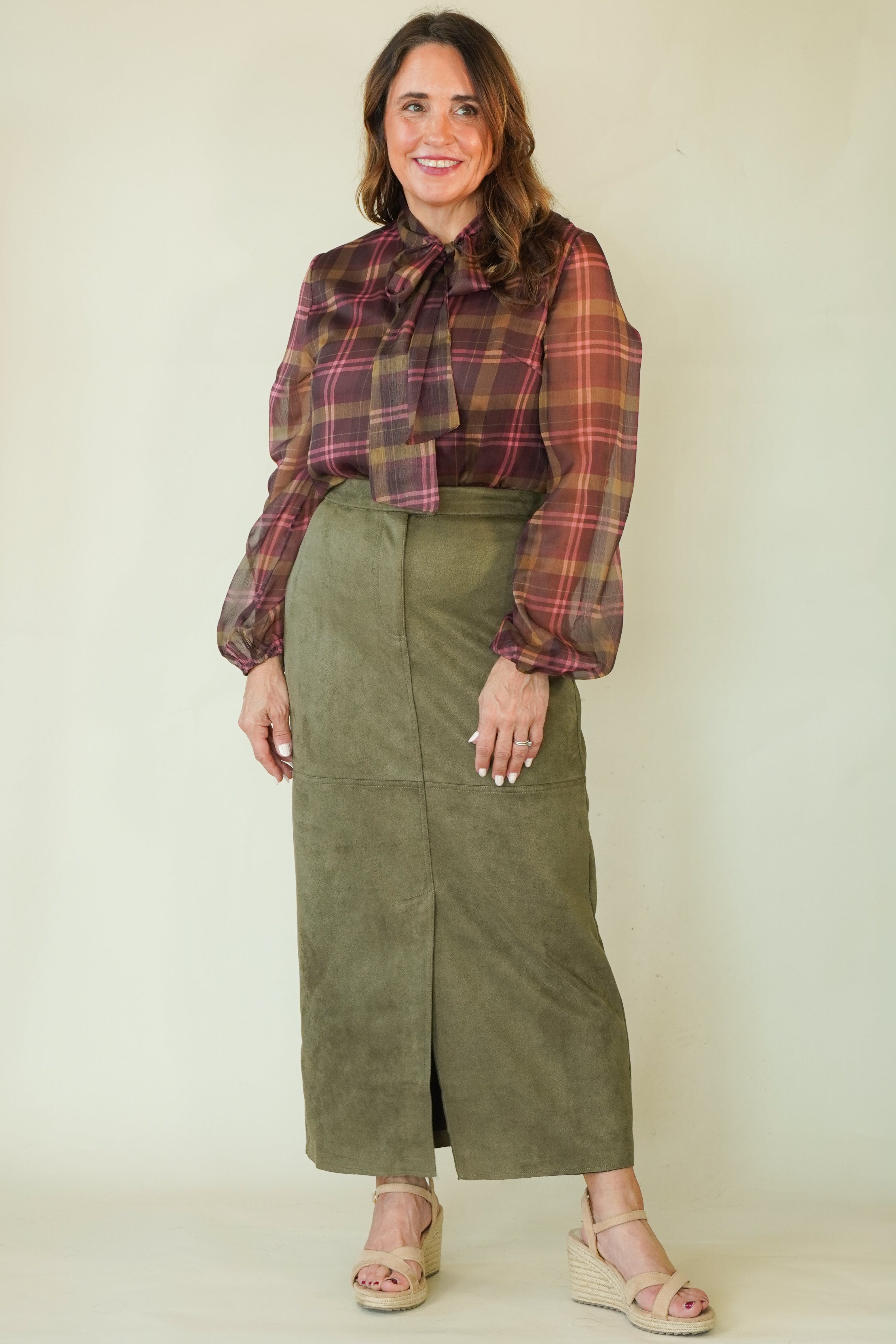 A woman modeling a long-sleeve olive green midi skirt paired with a brown and red plaid blouse, standing against a plain light beige studio background.