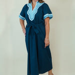 A woman modeling a dark teal blue maxi dress with light blue and white embroidered details on the V-neckline and sleeve cuffs, paired with white slide sandals, set against a plain cream background.