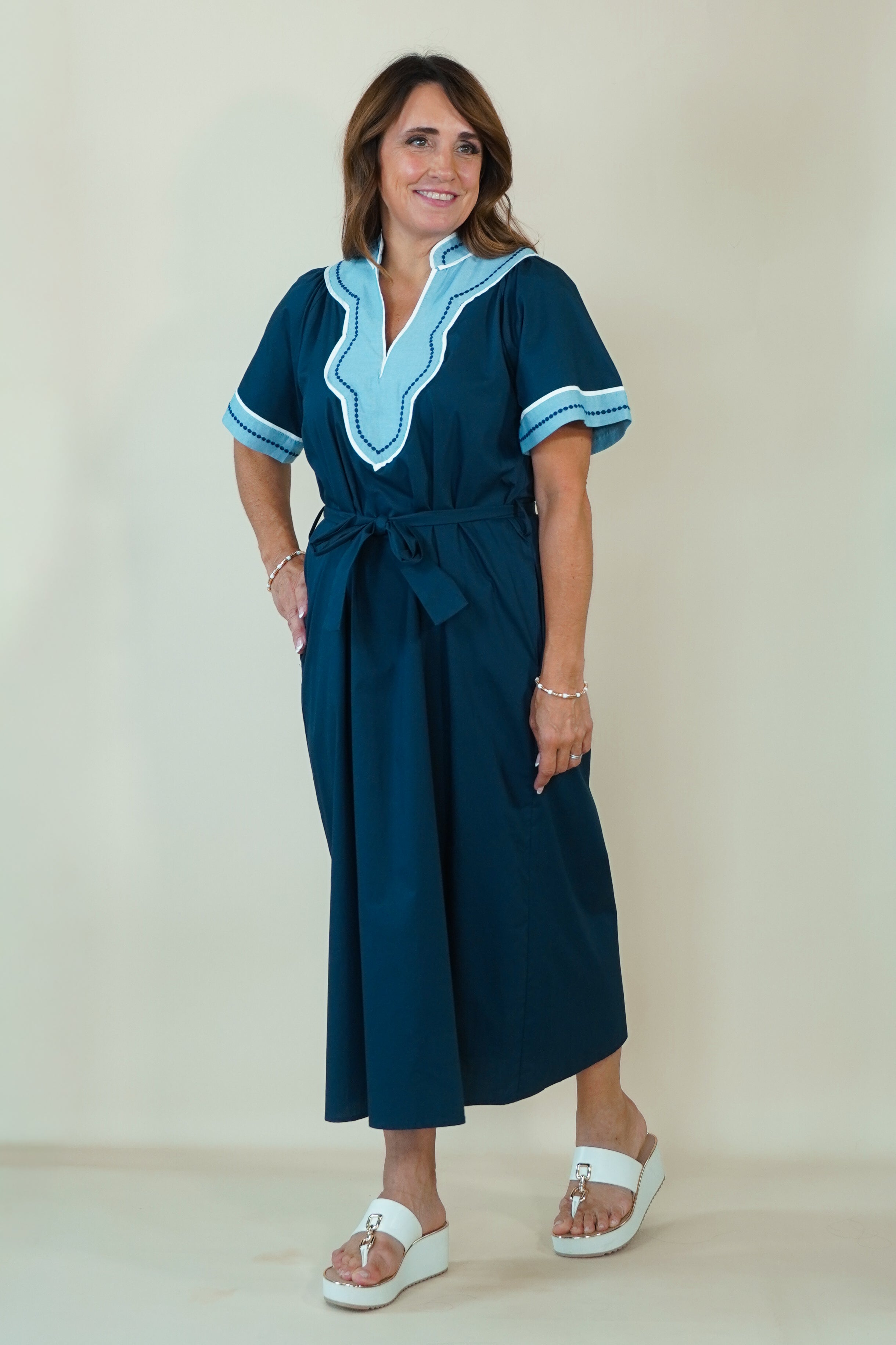 A woman modeling a dark teal blue maxi dress with light blue and white embroidered details on the V-neckline and sleeve cuffs, paired with white slide sandals, set against a plain cream background.