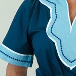 A woman modeling a dark teal blue maxi dress with light blue and white embroidered details on the V-neckline and sleeve cuffs, paired with white slide sandals, set against a plain cream background.