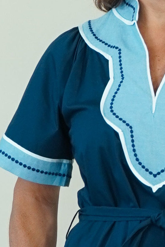 A woman modeling a dark teal blue maxi dress with light blue and white embroidered details on the V-neckline and sleeve cuffs, paired with white slide sandals, set against a plain cream background.