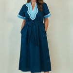 A woman modeling a dark teal blue maxi dress with light blue and white embroidered details on the V-neckline and sleeve cuffs, paired with white slide sandals, set against a plain cream background.