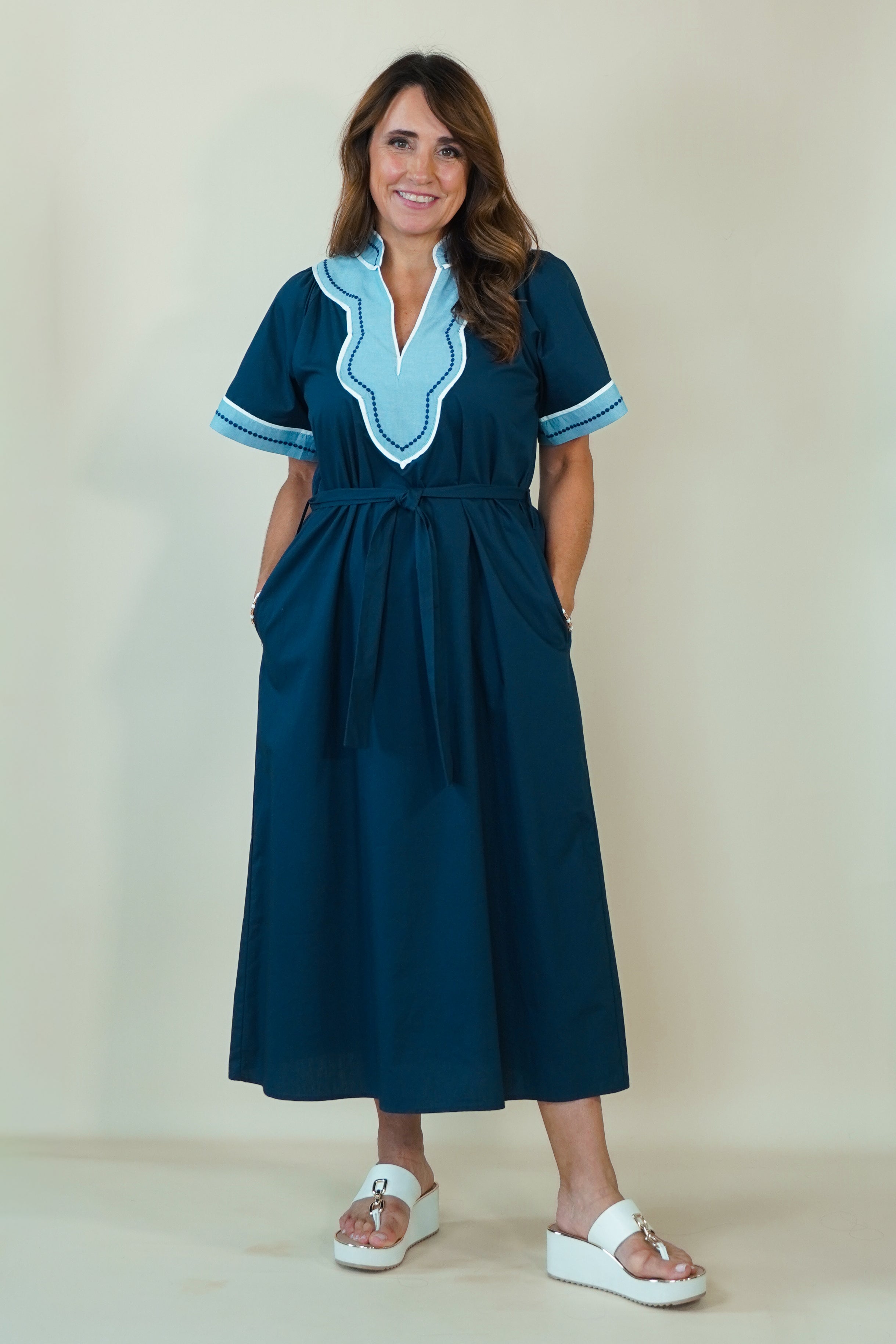 A woman modeling a dark teal blue maxi dress with light blue and white embroidered details on the V-neckline and sleeve cuffs, paired with white slide sandals, set against a plain cream background.