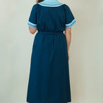 A woman modeling a dark teal blue maxi dress with light blue and white embroidered details on the V-neckline and sleeve cuffs, paired with white slide sandals, set against a plain cream background.