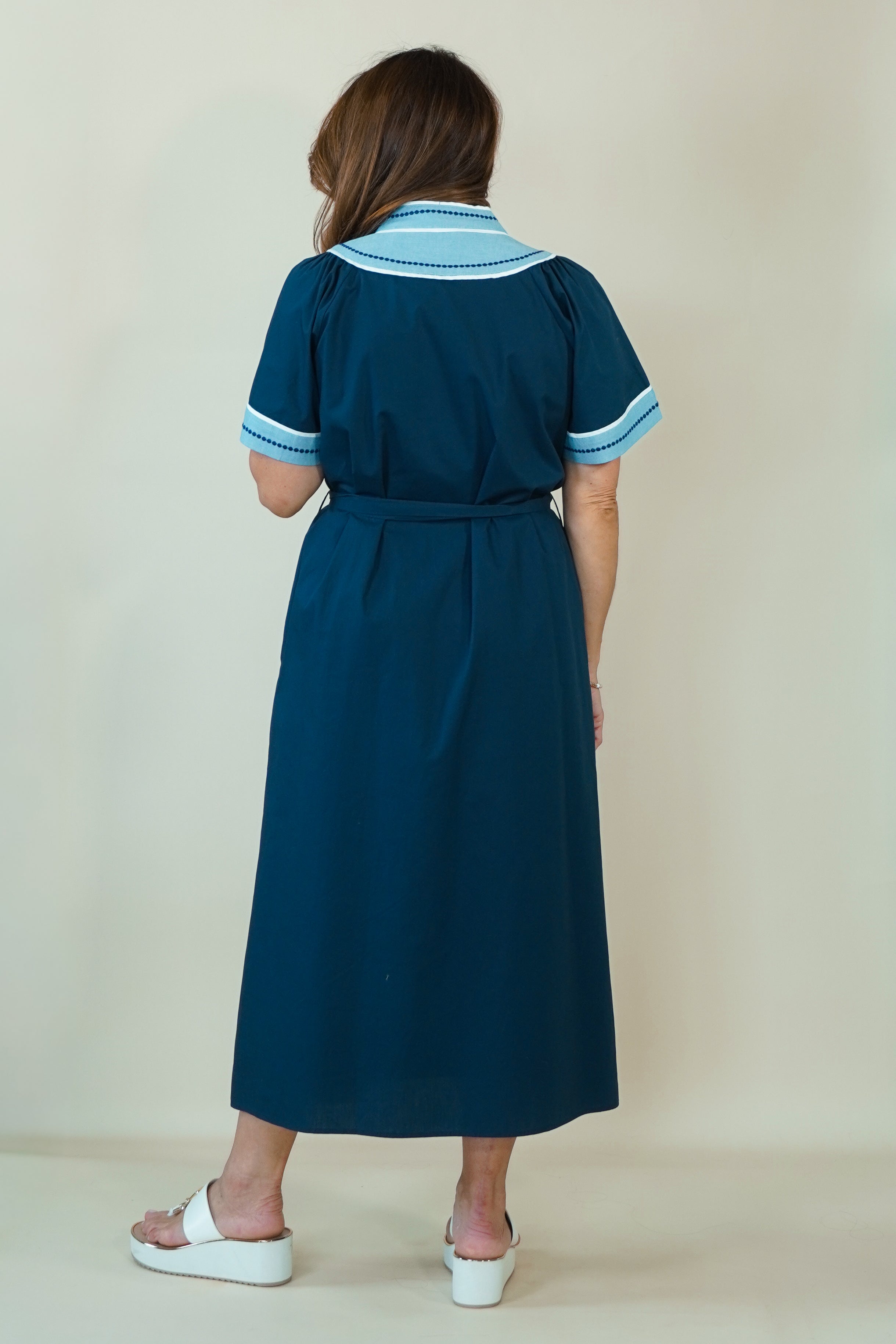 A woman modeling a dark teal blue maxi dress with light blue and white embroidered details on the V-neckline and sleeve cuffs, paired with white slide sandals, set against a plain cream background.