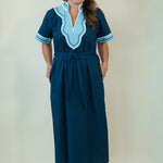A woman modeling a dark teal blue maxi dress with light blue and white embroidered details on the V-neckline and sleeve cuffs, paired with white slide sandals, set against a plain cream background.