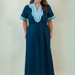 A woman modeling a dark teal blue maxi dress with light blue and white embroidered details on the V-neckline and sleeve cuffs, paired with white slide sandals, set against a plain cream background.