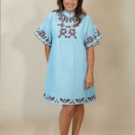 A woman modeling a light blue cotton dress with brown floral embroidery on the chest and hem, standing against a plain beige studio backdrop.