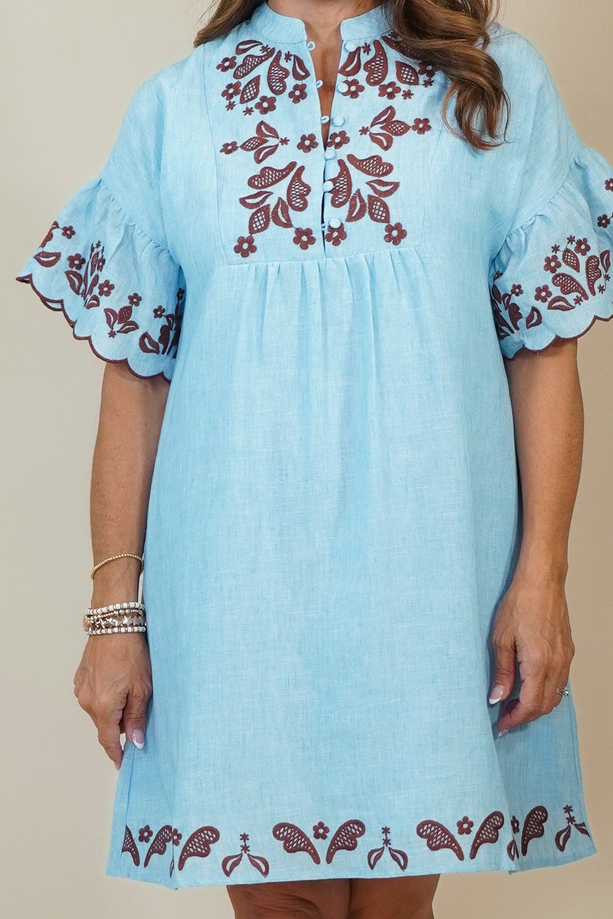 A woman modeling a light blue cotton dress with brown floral embroidery on the neckline, sleeve cuffs, and hem, standing against a neutral beige studio background.