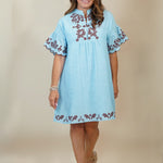 A woman modeling a light blue cotton tunic dress with brown floral embroidery on the chest, neckline, and hem, set against a plain beige studio background.