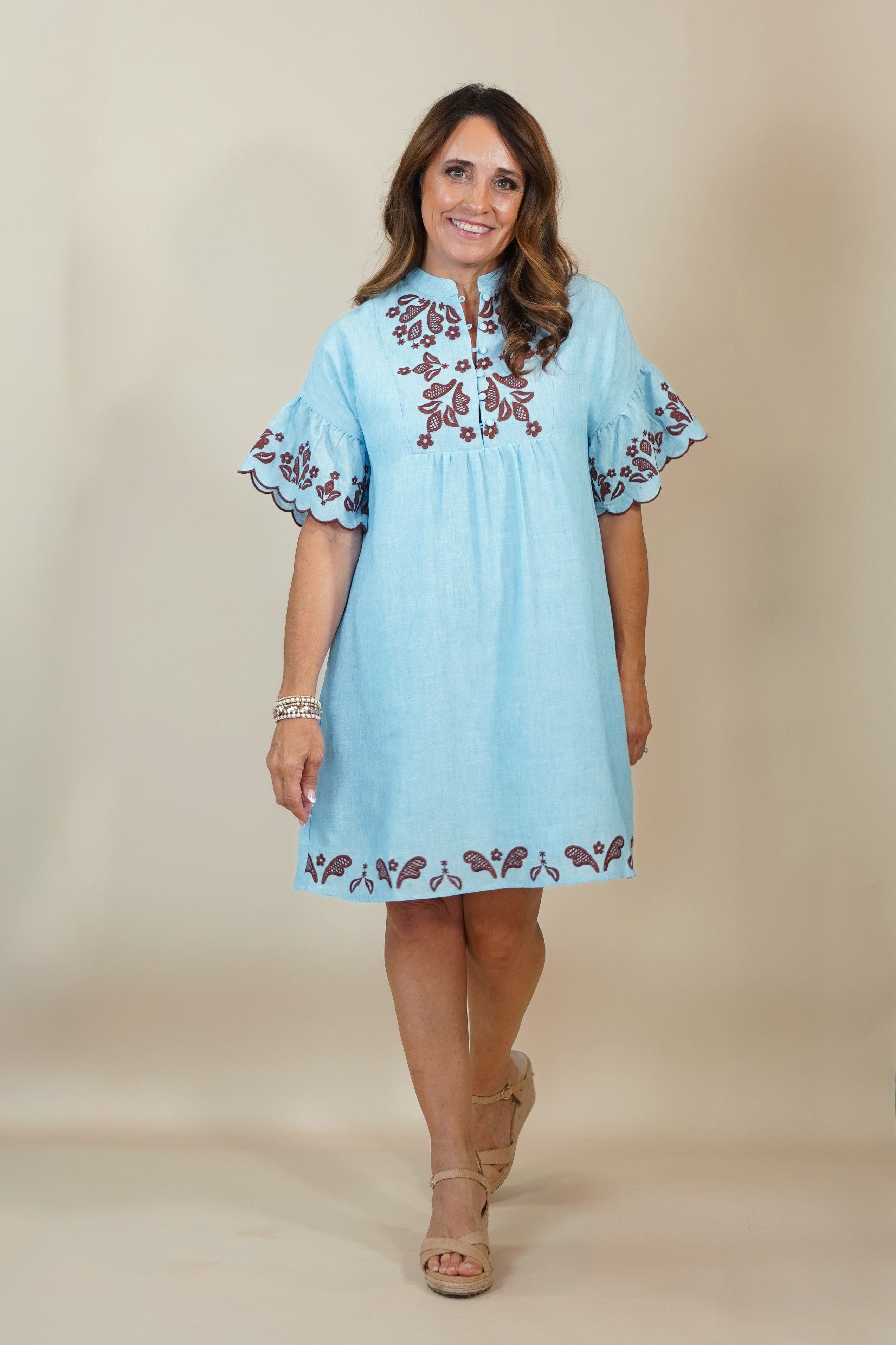 A woman modeling a light blue cotton tunic dress with brown floral embroidery on the chest, neckline, and hem, set against a plain beige studio background.