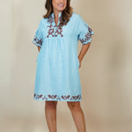 A woman modeling a light blue cotton tunic dress with brown and white floral embroidery on the neckline, sleeve cuffs, and hem, set against a plain beige studio background.