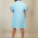 A woman viewed from the back wearing a light blue short-sleeve dress with ruffled sleeves and dark floral embroidery on the cuffs, paired with beige wedge sandals, against a plain beige studio backdrop.