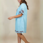 A woman modeling a light blue cotton tunic dress with brown floral embroidery on the sleeves and hem, paired with beige wedge sandals, set against a plain beige studio background.