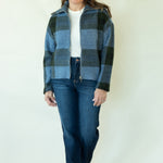A woman modeling a blue and black plaid zip-up jacket over a white t-shirt and dark blue jeans, standing against a plain off-white background.