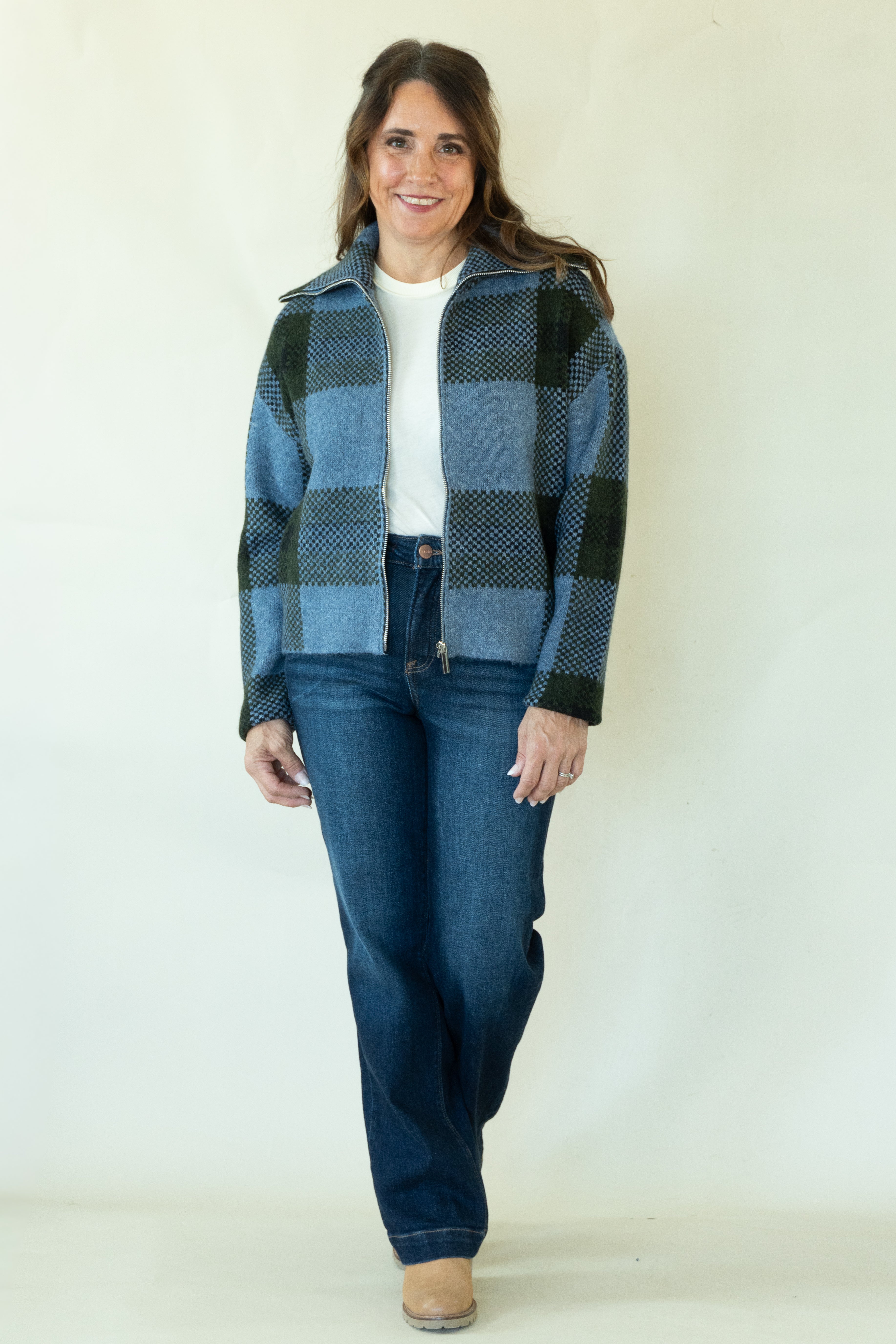 A woman modeling a blue and black plaid zip-up jacket over a white t-shirt and dark blue jeans, standing against a plain off-white background.