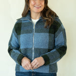A woman modeling a blue and black buffalo plaid zip-up fleece jacket with dark blue jeans and tan boots, standing against a plain off-white studio background.