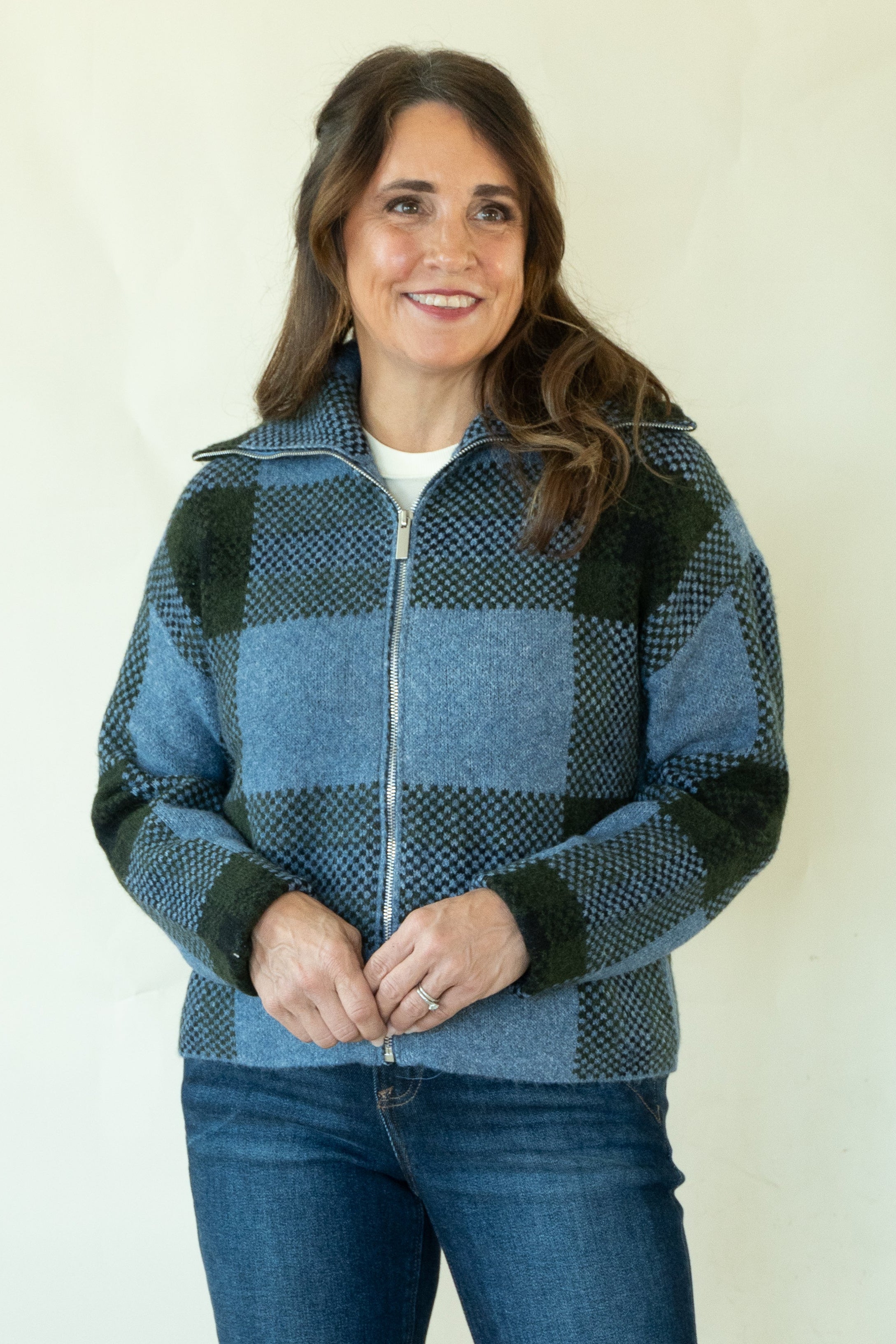 A woman modeling a blue and black buffalo plaid zip-up fleece jacket with dark blue jeans and tan boots, standing against a plain off-white studio background.